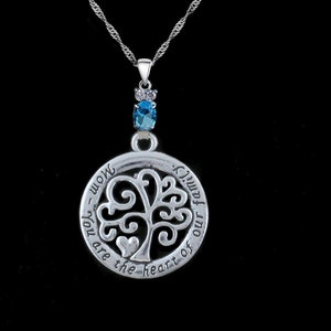 Peut inclure: Collier pendentif argenté avec un motif d'arbre de vie dans un cadre circulaire. Le pendentif porte l'inscription "Maman - Tu es le cœur de notre famille". Une pierre bleue et une chaîne délicate complètent le collier.
