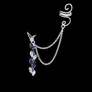 Op de afbeelding: Zilveren oormanchet met een hangende ketting en blauwe en witte kristallen accenten.