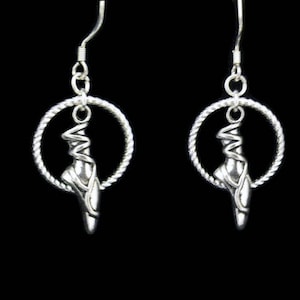 Könnte beinhalten: Silberne Ohrringe mit einem Ballerina-Schuh-Anhänger. Der Schuh ist mit einem Band verziert und hängt an einem silbernen Kreis.