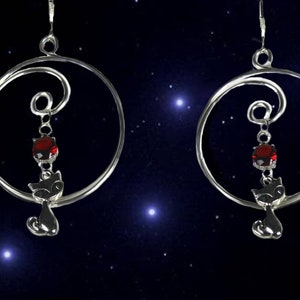Peut inclure: Une paire de boucles d'oreilles en argent en forme de cercle avec une pierre précieuse rouge pendante et un charme de chat en argent. Les boucles d'oreilles sont suspendues à un fil d'argent torsadé en forme de spirale.