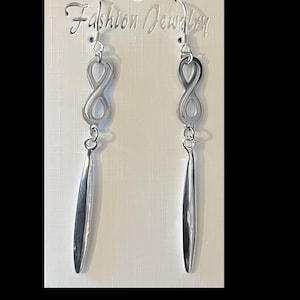 Puede incluir: Pendientes de plata con símbolo de infinito y una larga y fina gota de plata. Los pendientes están sobre un fondo blanco con el texto "Fashion Jewelry" impreso en la tarjeta.
