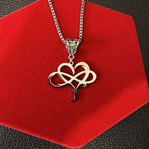 Op de afbeelding: Een zilverkleurige metalen ketting met een hanger die een hartvorm combineert met een oneindigheidssymbool. De hanger hangt aan een box-ketting en is voorzien van een decoratief filigraan-stijl oogje aan de bovenkant met een gepolijste afwerking.