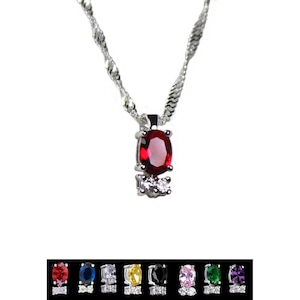 Op de afbeelding: Een zilveren ketting met een hanger met een rode ovale edelsteen omringd door twee kleinere heldere edelstenen.