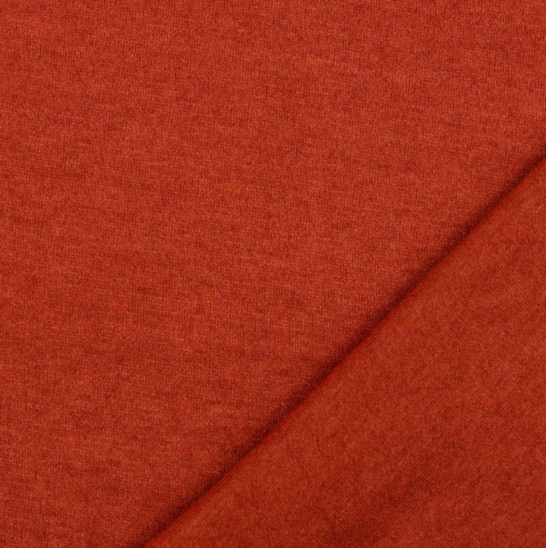 May include: Close-up of a textured, rust-colored fabric. The fabric is folded diagonally, creating a contrast in texture and shadow. The material appears soft and possibly suitable for clothing or home decor.