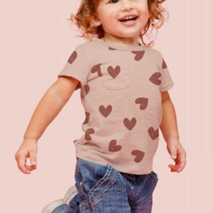May include: A young child wearing a light brown t-shirt with a brown heart pattern and blue jeans. The child is kneeling on a pink surface.