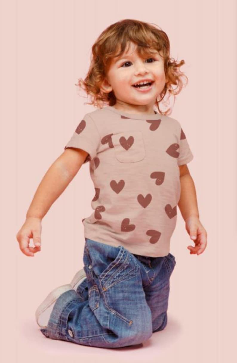May include: A young child wearing a light brown t-shirt with a brown heart pattern and blue jeans. The child is kneeling on a pink surface.