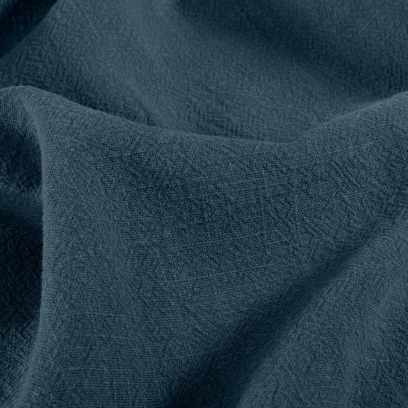May include: Close-up of a textured, dark teal fabric. The material is folded, showing the weave and subtle variations in color. The fabric appears soft and possibly linen or a similar natural fiber.