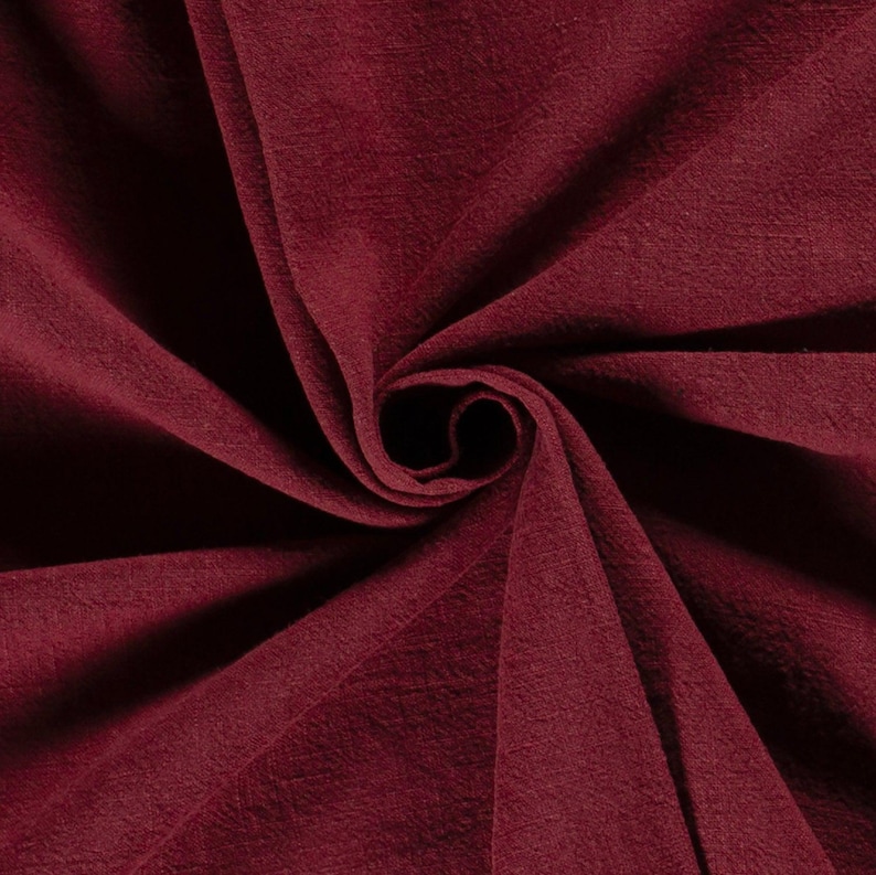 May include: Close-up of a deep burgundy fabric with a soft, textured appearance. The material is gathered and swirled, creating a spiral effect. The color is a rich, dark red, with subtle variations in tone.