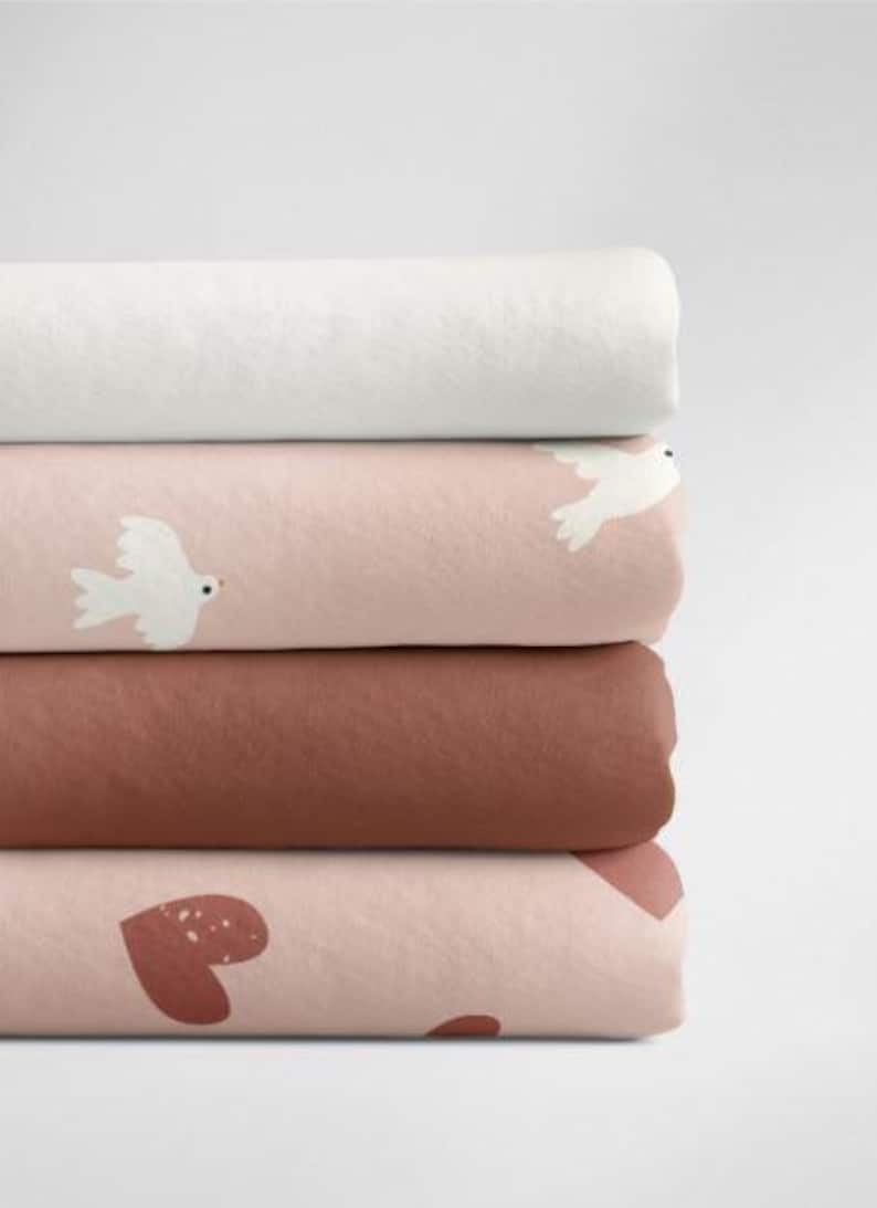May include: A stack of four fabric swatches in different colors and patterns. The top swatch is white, the second is pink with white doves, the third is brown, and the bottom is pink with brown hearts.
