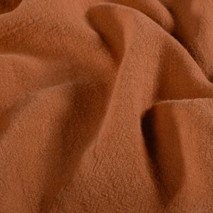 May include: Close-up of a textured, rust-colored fabric with soft folds and subtle variations in tone. The material appears to be a woven textile, possibly linen or a similar natural fiber, with a slightly rough texture.