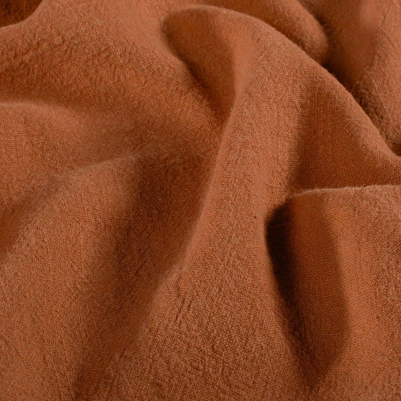 May include: Close-up of a textured, rust-colored fabric with soft folds and subtle variations in tone. The material appears to be a woven textile, possibly linen or a similar natural fiber, with a slightly rough texture.