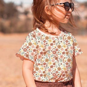 K&ouml;nnte beinhalten: Ein junges M&auml;dchen tr&auml;gt ein wei&szlig;es T-Shirt mit Blumenmuster und einen braunen Rock. Sie tr&auml;gt eine schwarze Sonnenbrille und hat lange braune Haare.