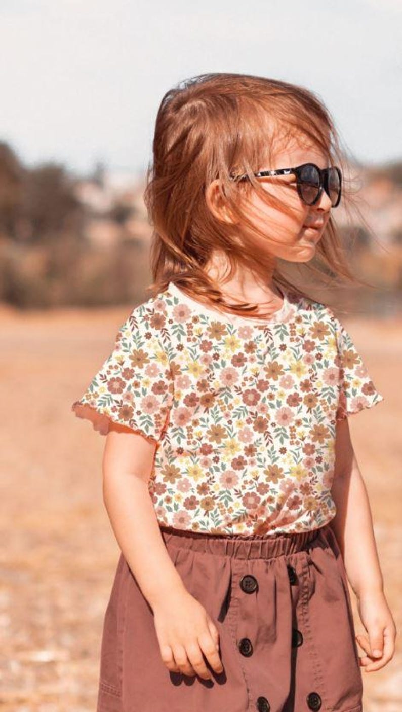 K&ouml;nnte beinhalten: Ein junges M&auml;dchen tr&auml;gt ein wei&szlig;es T-Shirt mit Blumenmuster und einen braunen Rock. Sie tr&auml;gt eine schwarze Sonnenbrille und hat lange braune Haare.