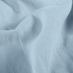 May include: Close-up of a light blue fabric with a textured weave. The fabric is soft and flowing, with subtle variations in color and texture. The image highlights the fabric's delicate appearance and drape.