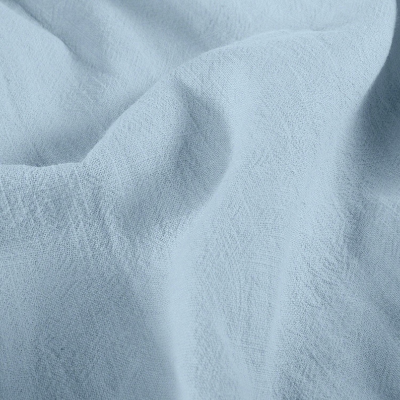 May include: Close-up of a light blue fabric with a textured weave. The fabric is soft and flowing, with subtle variations in color and texture. The image highlights the fabric's delicate appearance and drape.