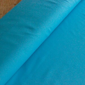 May include: A close-up of a turquoise blue fabric with a subtle speckled texture. The fabric is folded in half, revealing the smooth, even surface.