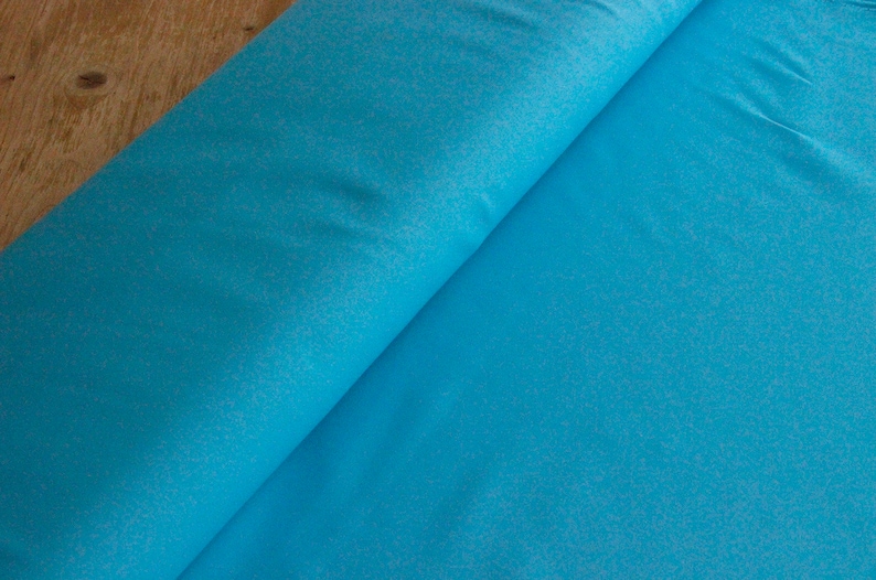 May include: A close-up of a turquoise blue fabric with a subtle speckled texture. The fabric is folded in half, revealing the smooth, even surface.