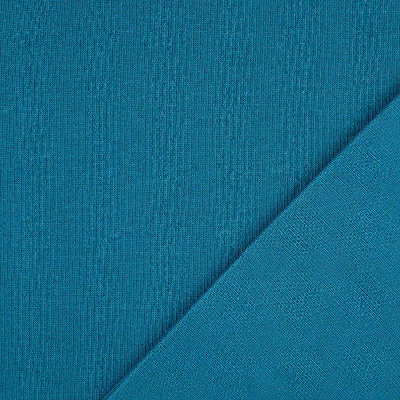 May include: Close-up of two pieces of teal fabric. The fabric on the left has a ribbed texture, while the fabric on the right is smooth. The two pieces are angled against each other, creating a diagonal line.