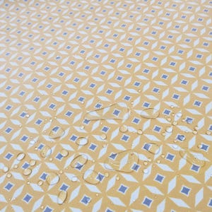 Könnte beinhalten: Nahaufnahme einer gelben, gemusterten Oberfläche mit einem sich wiederholenden geometrischen Design in Weiß und Blau. Wassertropfen sind über die Oberfläche verstreut, was auf ein wasserdichtes oder wasserabweisendes Material hindeutet. Das Muster ist eine Mischung aus Rauten und Quadraten.