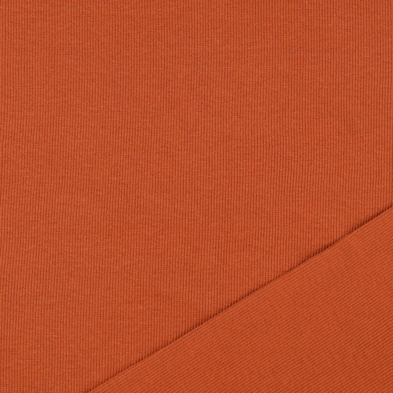 May include: Close-up of a textured, burnt orange fabric. The fabric has a ribbed pattern, with a diagonal section of the same material. The color is a warm, earthy tone, suitable for apparel or home decor.