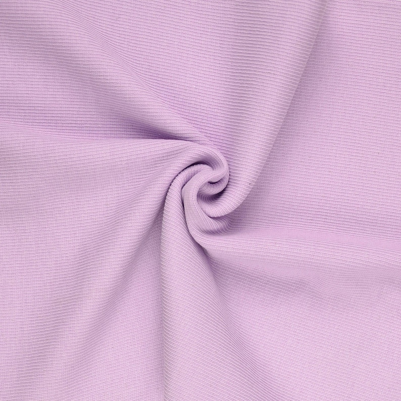May include: A close-up shot of a lavender fabric with a ribbed texture. The fabric is gathered and swirled, creating a soft, flowing appearance. The color is a light, pastel purple, and the fabric appears to be a knit or similar material.