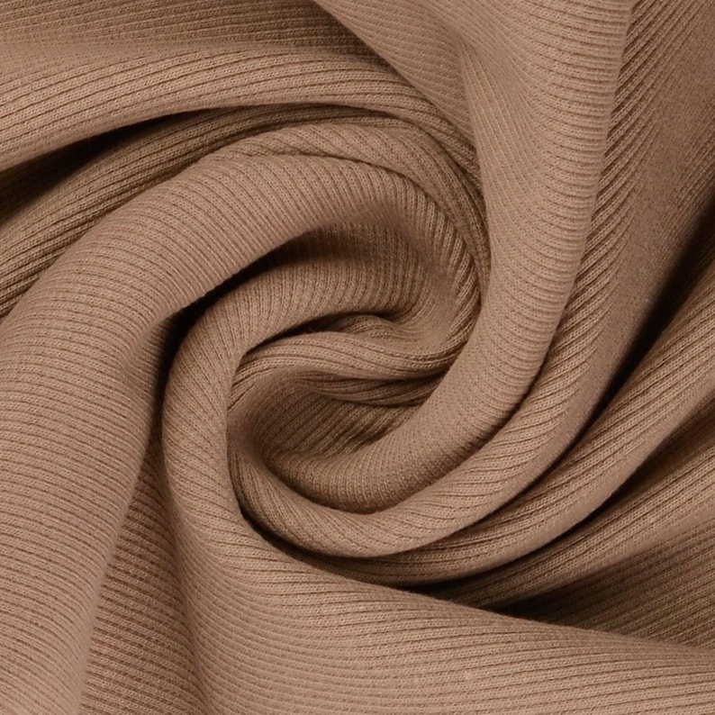 May include: Close-up of a folded, ribbed fabric in a warm, neutral brown color. The fabric has a soft texture with vertical ridges, creating a subtle pattern. The folds and shadows add depth to the image, highlighting the material's drape.