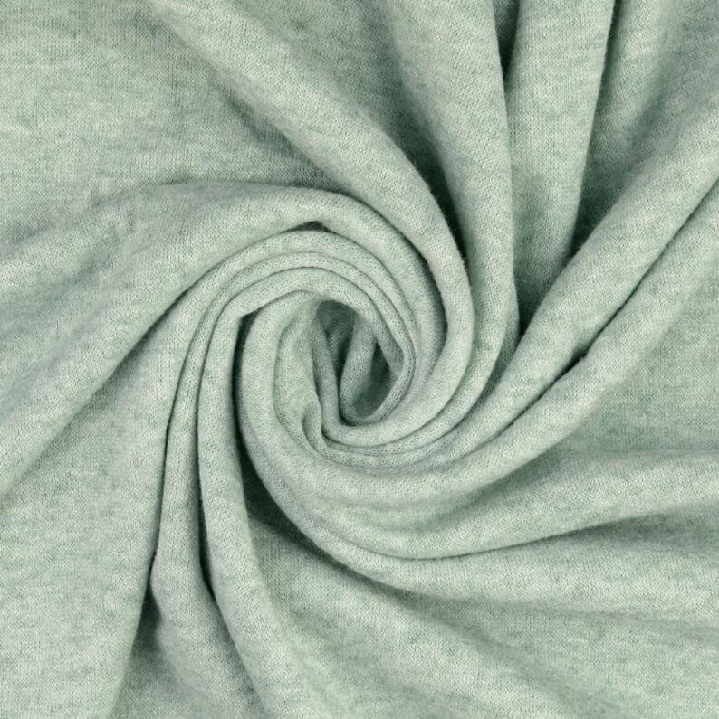 May include: Close-up of a soft, light green fabric with a swirling pattern. The fabric appears to be knit or woven, with a subtle texture and a muted, pastel color. The folds create shadows and depth.
