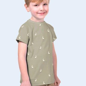 May include: A young child wearing a short-sleeved, olive green t-shirt with a pattern of small, white, crescent-shaped designs. The child is also wearing khaki-colored pants. The background is a light blue.