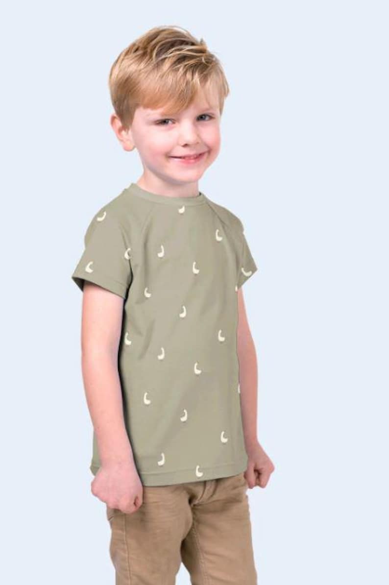 May include: A young child wearing a short-sleeved, olive green t-shirt with a pattern of small, white, crescent-shaped designs. The child is also wearing khaki-colored pants. The background is a light blue.