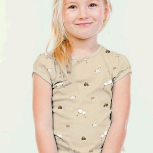 May include: A tan short-sleeved t-shirt featuring a pattern of white sheep, brown tractors, and brown fences. The shirt is worn by a young girl with blonde hair and blue jeans.