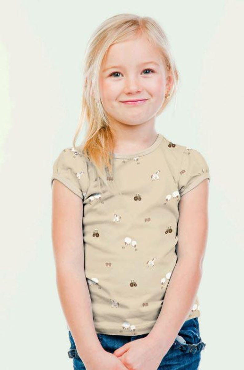May include: A tan short-sleeved t-shirt featuring a pattern of white sheep, brown tractors, and brown fences. The shirt is worn by a young girl with blonde hair and blue jeans.