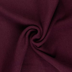 May include: Close-up of a swatch of burgundy fabric. The fabric has a ribbed texture and is draped in a swirling pattern. The color is a deep, rich shade of red-purple.