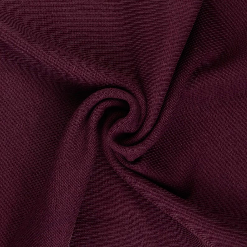 May include: Close-up of a swatch of burgundy fabric. The fabric has a ribbed texture and is draped in a swirling pattern. The color is a deep, rich shade of red-purple.