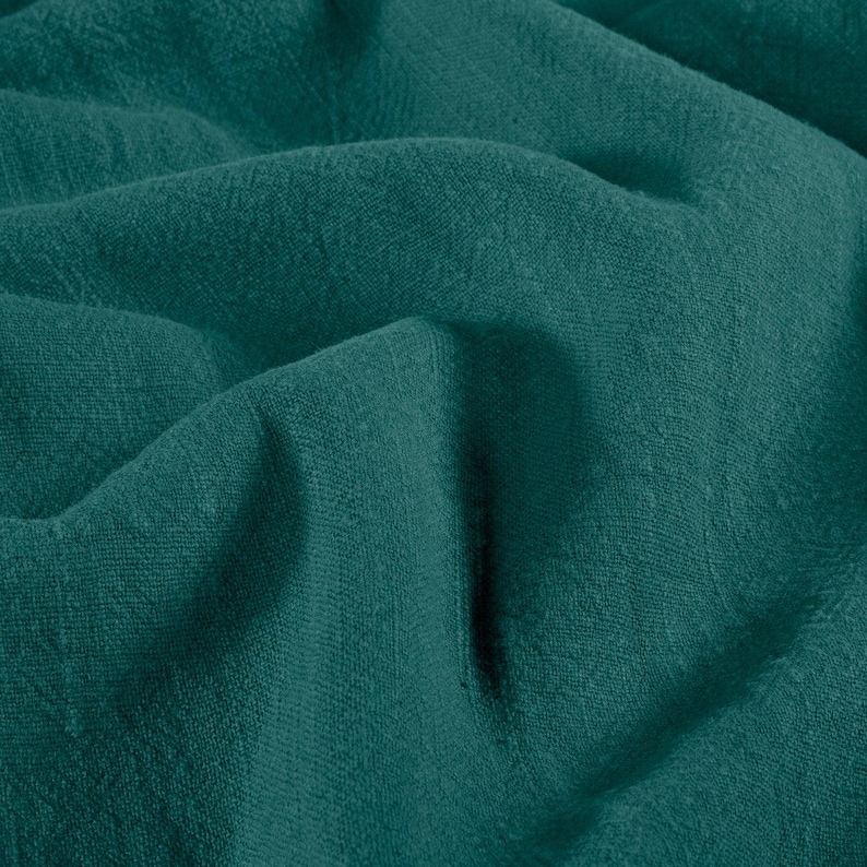May include: Close-up of a teal-colored fabric with a textured weave. The fabric is draped, creating soft folds and shadows. The color is a deep, saturated teal, with subtle variations in tone across the surface. The material appears to be lightweight and breathable.