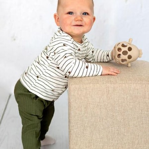 May include: A baby wearing a striped long-sleeve shirt and olive green pants. The baby is holding a beige toy and leaning on a beige ottoman. The outfit includes white socks. The background is a white wall and a light wood floor.