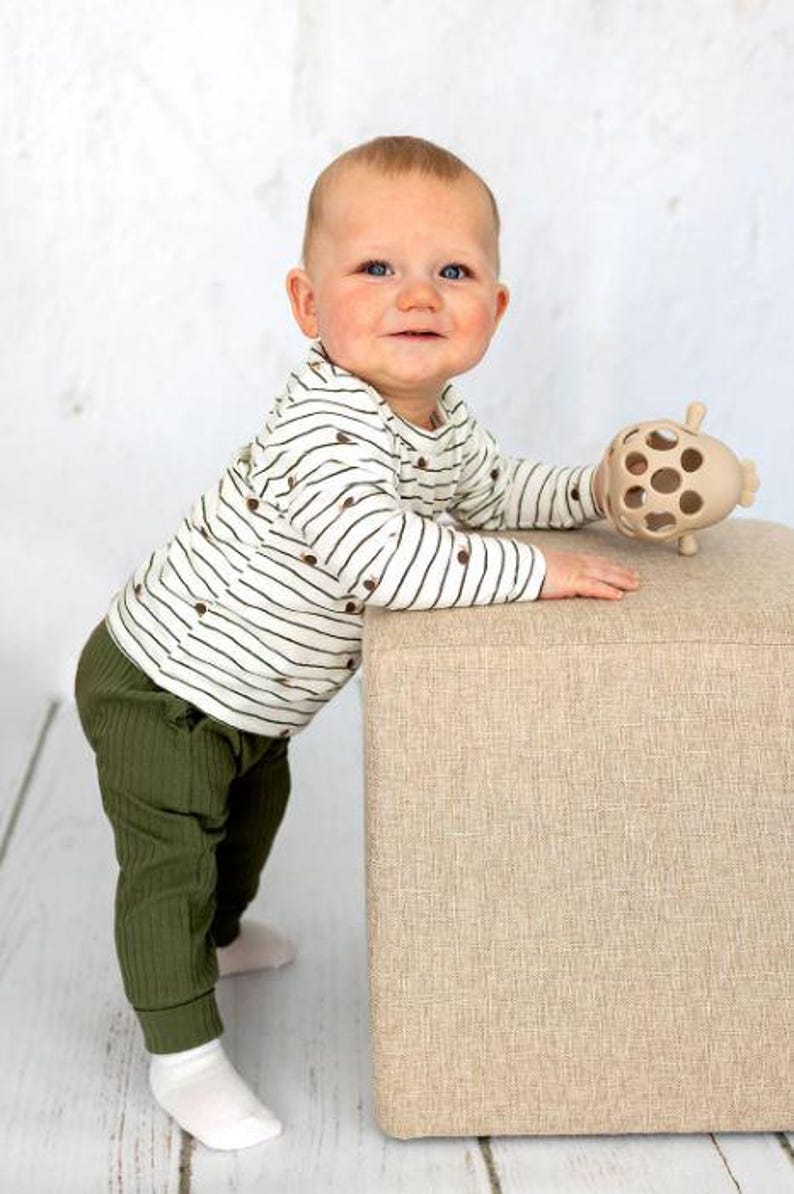 May include: A baby wearing a striped long-sleeve shirt and olive green pants. The baby is holding a beige toy and leaning on a beige ottoman. The outfit includes white socks. The background is a white wall and a light wood floor.
