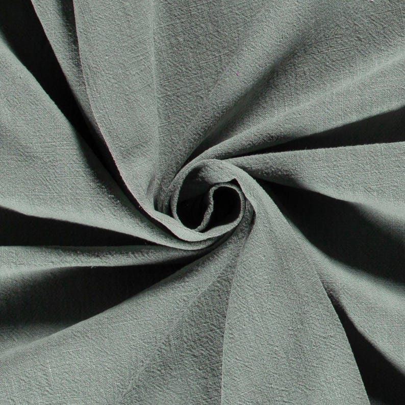 May include: Close-up of a swirl of sage green fabric. The fabric has a textured appearance with visible woven threads. The folds create shadows and highlights, emphasizing the material's softness and drape. The fabric is likely used for clothing or home decor.