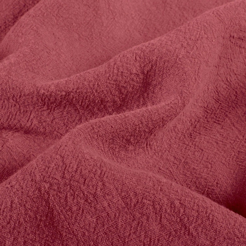 May include: Close-up of a textured, crumpled fabric in a deep rose color. The fabric has a slightly rough, woven appearance with visible folds and shadows, suggesting a soft, draping material. The overall effect is one of warmth and depth.