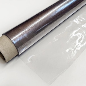 Könnte beinhalten: Eine Rolle transparente Kunststofffolie mit einem metallisch-silbernen Kern. Die Kunststofffolie ist teilweise abgerollt und zeigt eine transparente Folie. Der Kern besteht aus Pappe.