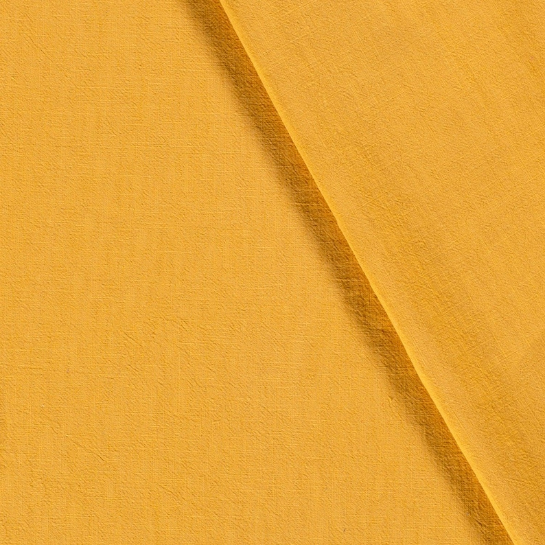 May include: Close-up of a folded piece of fabric in a warm, golden-yellow hue. The material has a subtle texture, with visible woven threads. The fabric is draped diagonally, creating a play of light and shadow.