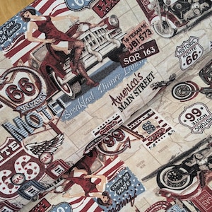 Op de afbeelding: Stof met een vintage Route 66-thema. Het ontwerp bevat illustraties van motorfietsen, klassieke auto's en pin-up girls, samen met verkeersborden en de tekst "America's Main Street". Het kleurenpalet omvat rood, wit, blauw en beige.