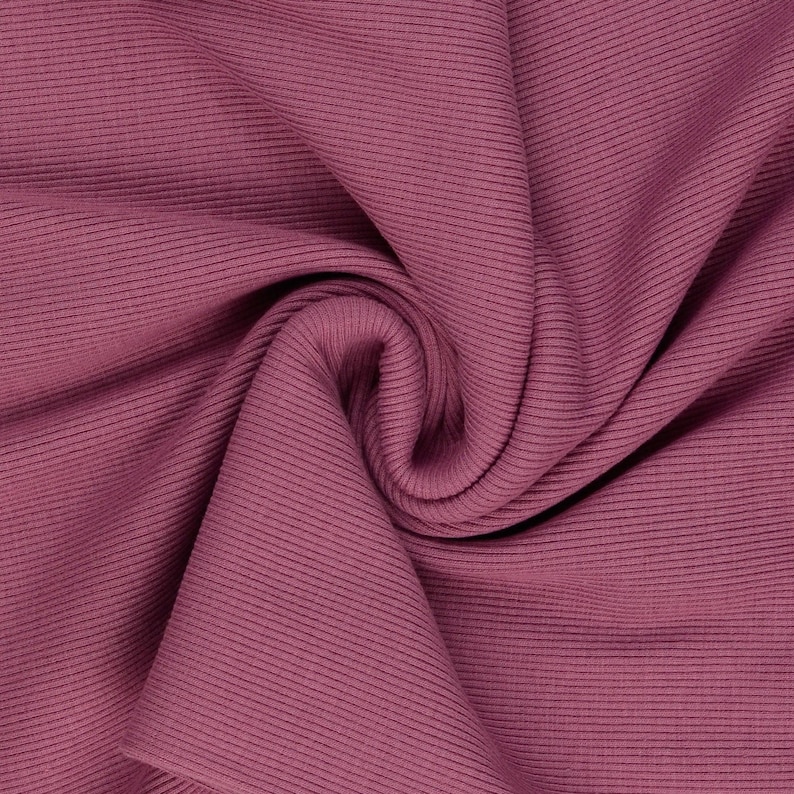May include: Close-up of a folded fabric in a deep rose color. The fabric has a ribbed texture with vertical lines running across the material. The folds create shadows and depth, highlighting the texture and color of the fabric.