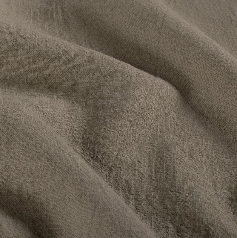 May include: Close-up of a textured, taupe-colored fabric. The material has a slightly wrinkled appearance, with visible woven threads and subtle variations in tone. The fabric drapes in soft folds, creating shadows and highlights.