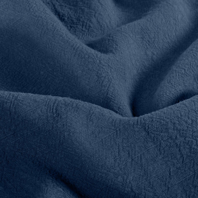 May include: Close-up of a textured, navy blue fabric. The fabric is folded and draped, creating shadows and highlighting the woven texture. The deep blue color suggests a natural dye.