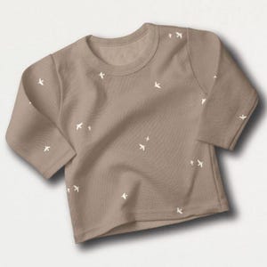 May include: A tan-colored long-sleeved shirt with a crew neck. The shirt is adorned with small, white bird-shaped designs scattered across the fabric. The garment is made of a soft material, suitable for casual wear.
