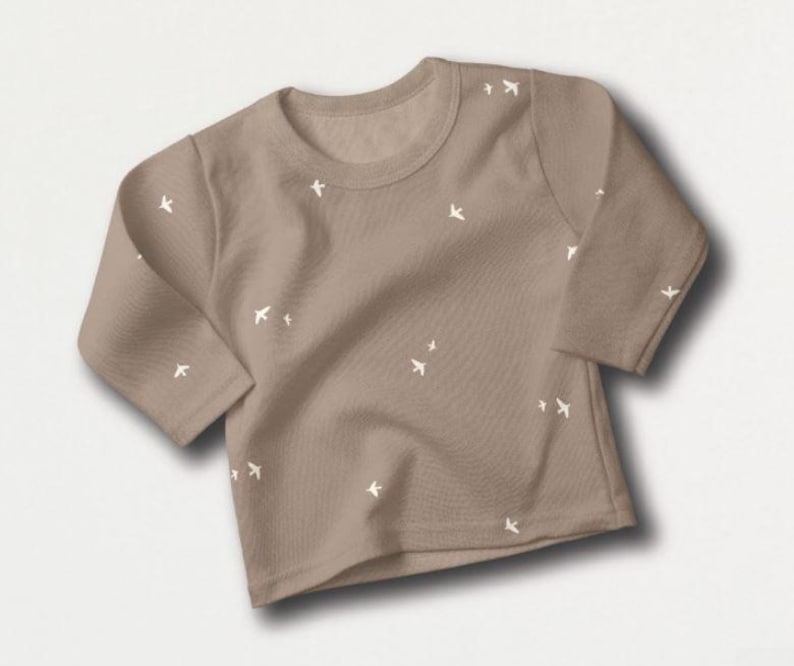 May include: A tan-colored long-sleeved shirt with a crew neck. The shirt is adorned with small, white bird-shaped designs scattered across the fabric. The garment is made of a soft material, suitable for casual wear.