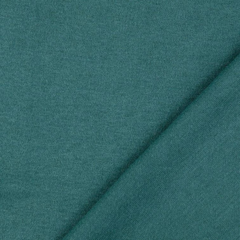 May include: Close-up of a folded piece of teal fabric. The fabric has a soft, textured appearance, with subtle variations in color. The material is draped diagonally across the frame, showcasing its drape and texture.