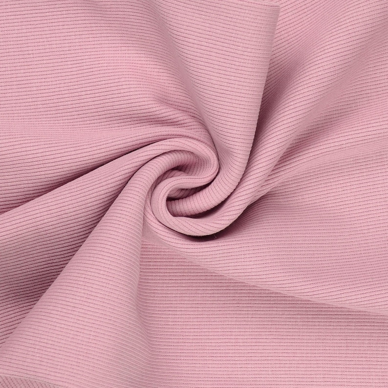 May include: Close-up of a swatch of pink ribbed fabric. The fabric is folded and draped, showing the texture and direction of the ribbing. The color is a soft, muted pink.
