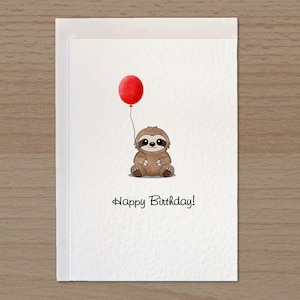 May include: A white greeting card featuring a brown cartoon sloth sitting and holding a single red balloon by a thin string. Below the sloth, the text Happy Birthday is printed in a simple black font. The card has a textured paper surface.