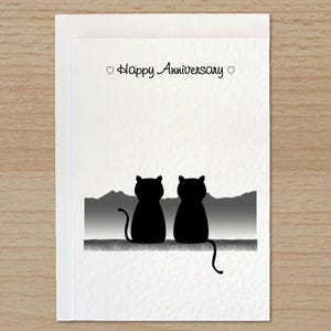 May include: A white greeting card featuring the black silhouette of two cats sitting side by side, facing a grey mountain landscape. The text "Happy Anniversary" is printed in black at the top, flanked by two small black heart symbols.